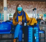 A woman wearing a face mask sits next to her blue suitcase in a travel lounge